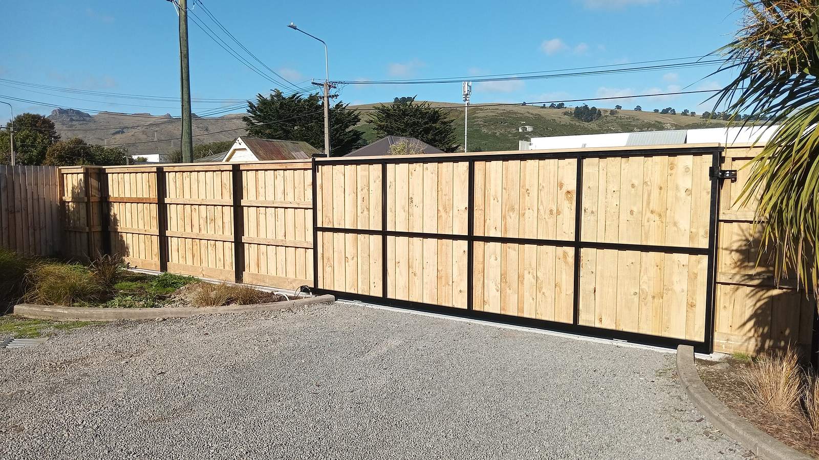 Timber paling fence and a black framed sliding gate across a gravel driveway, with the Port Hills behind