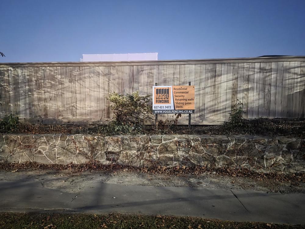 Vogel Fencing sign board fixed to a tall timber paling fence built along the top of a stone retaining wall
